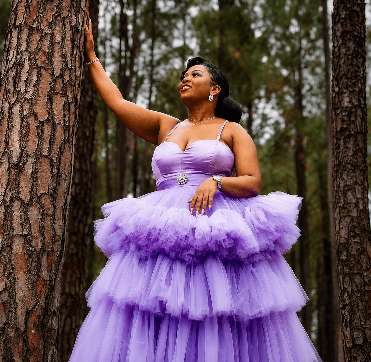 A woman in a layered lavender tulle gown standing among pine trees in a forest, one hand resting on a tree trunk