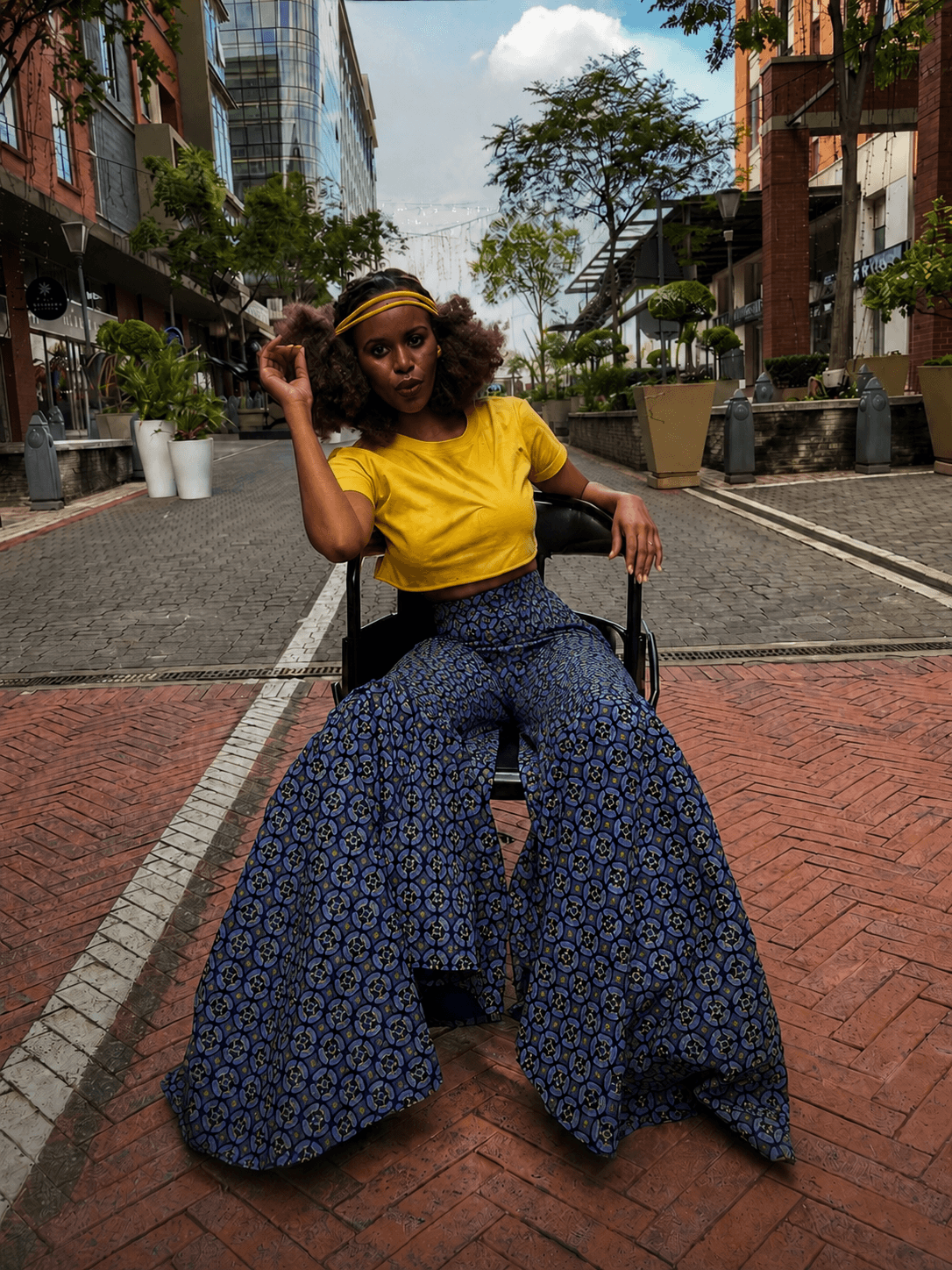 A woman wearing a cropped yellow top and wide-leg blue print palazzo pants, seated on a paved city street lined with shops and trees